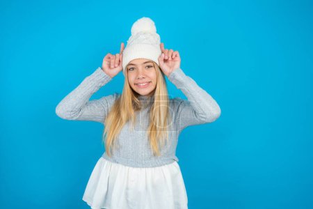 Téléchargez les photos : Funny belle enfant fille portant chapeau tricoté blanc et pull bleu montre cornes, doigts sur le geste de la tête, posant stupide et mignon - en image libre de droit