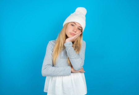 Photo for Very bored Beautiful kid girl wearing white knitted hat and blue sweater holding hand on cheek while support it with another crossed hand, looking tired and sick, - Royalty Free Image
