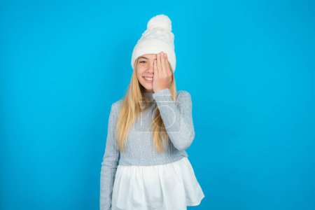 Téléchargez les photos : Belle gamine portant un chapeau tricoté blanc et un pull bleu couvrant un œil avec sa main, sourire confiant sur le visage et émotion surprise. - en image libre de droit