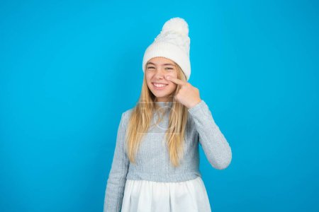 Beautiful kid girl wearing white knitted hat and blue sweater pointing unhappy to pimple on forehead, ugly infection of blackhead. Acne and skin problem