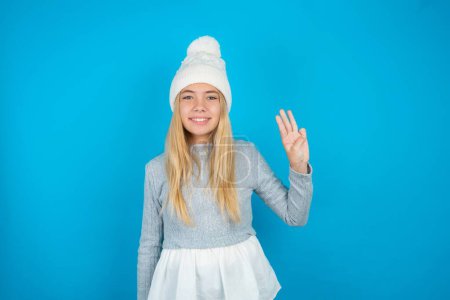 Photo for Beautiful kid girl wearing white knitted hat and blue sweater showing and pointing up with fingers number three while smiling confident and happy. - Royalty Free Image