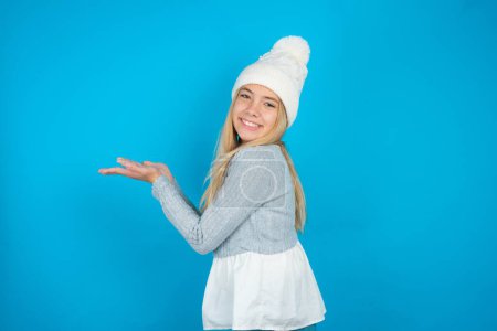 Téléchargez les photos : Belle fille enfant portant chapeau tricoté blanc et pull bleu pointant de côté avec les mains ouvertes paumes montrant l'espace de copie, présentant la publicité souriant heureux excité - en image libre de droit