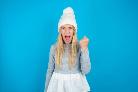 Beautiful kid girl wearing white knitted hat and blue sweater angry and mad raising fist frustrated and furious while shouting with anger. Rage and aggressive concept.