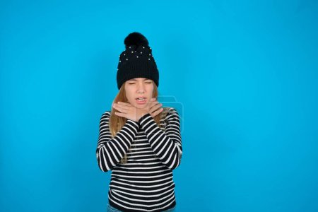 Photo for Beautiful kid girl wearing knitted black hat and striped turtleneck over blue background shouting suffocate because painful strangle. Health problem. Asphyxiate and suicide concept. - Royalty Free Image