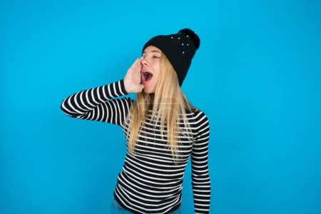 Photo for Beautiful kid girl wearing knitted black hat and striped turtleneck over blue background shouting and screaming loud to side with hand on mouth. Communication concept. - Royalty Free Image