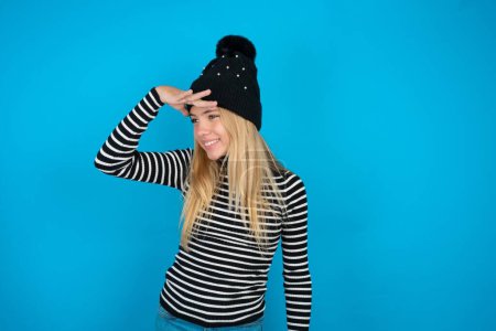 Photo for Beautiful kid girl wearing knitted black hat and striped turtleneck over blue background very happy and smiling looking far away with hand over head. Searching concept. - Royalty Free Image