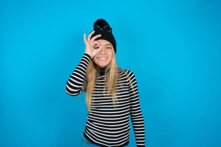 Téléchargez les photos : Belle enfant fille portant bonnet noir tricoté et col roulé rayé sur fond bleu faisant un geste correct avec la main souriant, oeil regardant à travers les doigts avec un visage heureux. - en image libre de droit