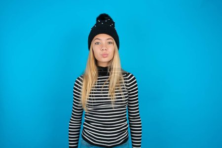 Shot of pleasant looking beautiful kid girl wearing knitted black hat and striped turtleneck over blue background pouts lips, looks at camera, Human facial expressions