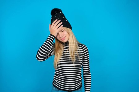 Photo for A very upset and lonely beautiful kid girl wearing knitted black hat and striped turtleneck over blue background crying - Royalty Free Image