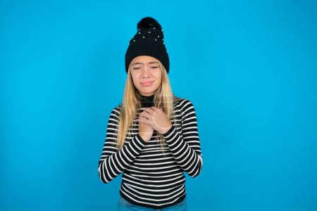 Photo for Sad beautiful kid girl wearing knitted black hat and striped turtleneck over blue background desperate and depressed with tears on her eyes suffering pain and depression  in sadness facial expression and emotion concept - Royalty Free Image