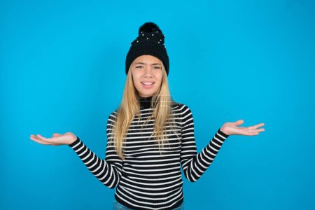 Indignant beautiful kid girl wearing knitted black hat and striped turtleneck over blue background gestures in bewilderment, frowns face with dissatisfaction.