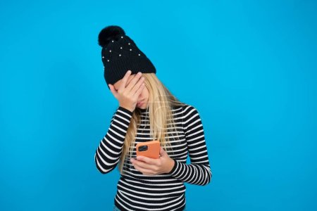 Photo for Beautiful kid girl wearing knitted black hat and striped turtleneck over blue background looking at smart phone feeling sad holding hand on face. - Royalty Free Image