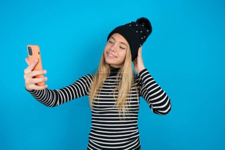 Téléchargez les photos : Belle enfant fille portant tricoté chapeau noir et col roulé rayé sur fond bleu souriant et prendre un selfie prêt à le poster sur ses médias sociaux. - en image libre de droit