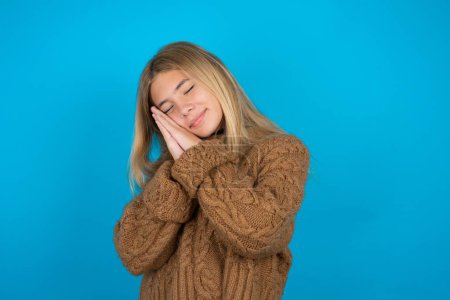 Photo for Beautiful kid girl wearing brown knitted sweater leans on pressed palms closes eyes and has pleasant smile dreams about something - Royalty Free Image
