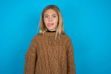 Photo for Funny Beautiful kid girl wearing brown knitted sweater makes grimace and crosses eyes plays fool has fun alone sticks out tongue. - Royalty Free Image