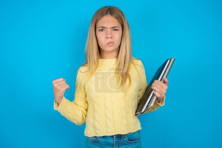 Photo for Irritated Beautiful kid girl wearing yellow sweater blows cheeks with anger and raises clenched fists expresses rage and aggressive emotions. Furious model - Royalty Free Image