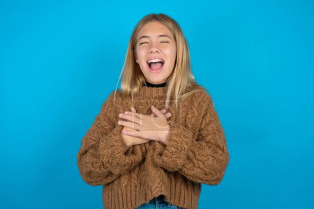 Foto de Hermosa niña usando suéter de punto marrón expresa felicidad, ríe agradablemente, mantiene las manos en el corazón - Imagen libre de derechos