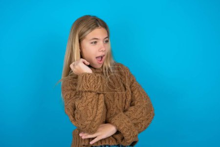 Foto de Sorprendida Hermosa niña usando suéter de punto marrón se ve a un lado sorprendentemente con la boca abierta. - Imagen libre de derechos