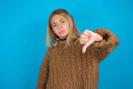 Foto de Hermosa niña usando suéter de punto marrón con aspecto infeliz y enojado mostrando rechazo y negativo con los pulgares hacia abajo gesto. Mala expresión. - Imagen libre de derechos