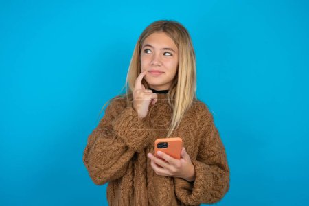 Photo for Beautiful kid girl wearing brown knitted sweater thinks deeply about something, uses modern mobile phone, tries to made up good message, keeps index finger near lips. - Royalty Free Image
