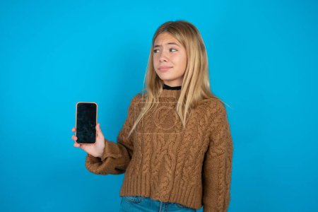 Téléchargez les photos : Belle fille enfant portant un pull en tricot brun tient un nouveau téléphone portable et semble mystérieux montre l'affichage vierge de cellulaire moderne - en image libre de droit