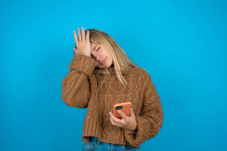 Photo for Upset depressed Beautiful kid girl wearing brown knitted sweater makes face palm as forgot about something important holds mobile phone expresses sorrow and regret blames - Royalty Free Image