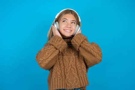 Photo for Joyful Beautiful kid girl wearing brown knitted sweater sings song keeps hand near mouth as if microphone listens favorite playlist via headphones - Royalty Free Image
