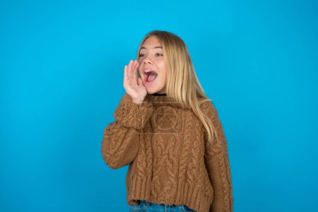 Foto de Hermosa niña usando suéter de punto marrón mirada espacio vacío sosteniendo la cara de la mano y gritando o llamando a alguien. - Imagen libre de derechos