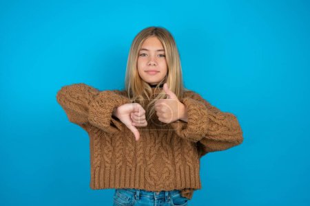 Photo for Beautiful kid girl wearing brown knitted sweater showing thumb up down sign - Royalty Free Image