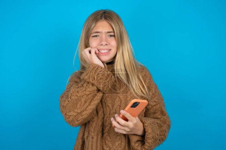 Photo for Afraid funny Beautiful kid girl wearing brown knitted sweater holding telephone and biting nails - Royalty Free Image