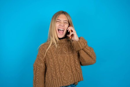 Overemotive happy Beautiful kid girl wearing brown knitted sweater laughs out positively hears funny story from friend during telephone conversation