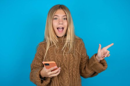 Photo for Astonished Beautiful kid girl wearing brown knitted sweater holding her telephone and pointing with finger aside at empty copy space - Royalty Free Image