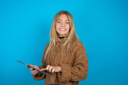 Foto de Foto de feliz alegre Hermosa niña usando marrón suéter de punto mantenga la tableta navegar por Internet - Imagen libre de derechos