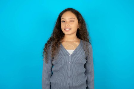 Coquettish Beautiful kid girl wearing casual jacket over blue background smiling happily, blinking at camera in a playful manner, flirting with you.