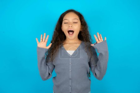 Photo for Emotive Beautiful kid girl wearing casual jacket over blue background laughs loudly, hears funny joke or story, raises palms with satisfaction, - Royalty Free Image
