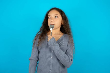 Very hungry Beautiful kid girl wearing casual jacket over blue background holding spoon into mouth dream of tasty meal