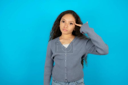 Photo for Unhappy Beautiful kid girl wearing casual jacket over blue background makes suicide gesture and imitates gun with hand, curves lips keeps two fingers on temple, shoots, being tired of everything, - Royalty Free Image
