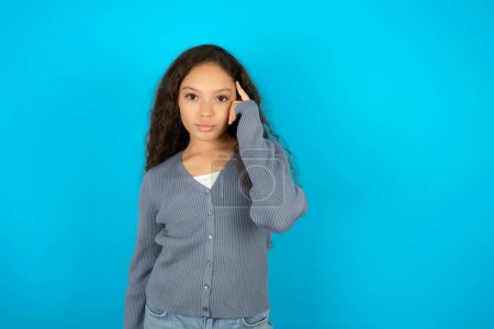 Photo for Beautiful kid girl wearing casual jacket over blue background tries to memorize something, keeps fore finger on temple, reminds information for exam, - Royalty Free Image