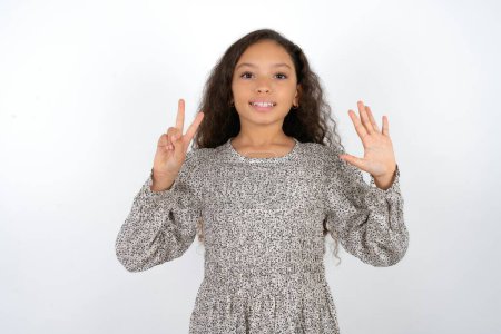 Foto de Hermosa adolescente vistiendo vestido gris sobre fondo blanco mostrando y apuntando hacia arriba con los dedos número siete mientras sonríe confiado y feliz. - Imagen libre de derechos