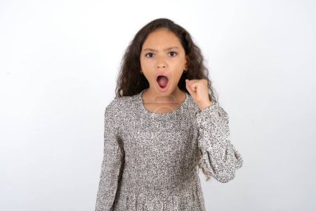 beautiful teen girl wearing grey dress over white background angry and mad raising fist frustrated and furious while shouting with anger. Rage and aggressive concept.