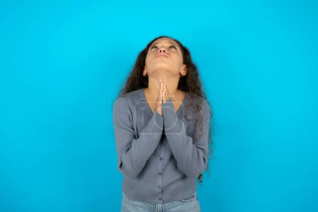 Téléchargez les photos : Belle jeune fille portant une veste décontractée sur fond bleu mendier et prier avec les mains ainsi que l'expression d'espoir sur le visage très émotionnel et inquiet. Mon Dieu ! - en image libre de droit