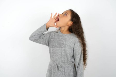 Foto de Hermosa chica adolescente con vestido gris sobre fondo blanco gritando y gritando fuerte a un lado con la mano en la boca. Concepto de comunicación. - Imagen libre de derechos