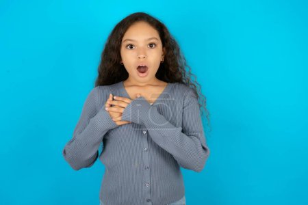 Scared Beautiful kid girl wearing casual jacket over blue background looks with frightened expression, keeps hands on chest, being puzzled to notice something strange, People, hush reaction and emotions.