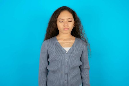 Photo for Dismal gloomy rejected Beautiful kid girl wearing casual jacket over blue background has problems and difficulties, curves lower lip and closes eyes in despair, being in depression - Royalty Free Image