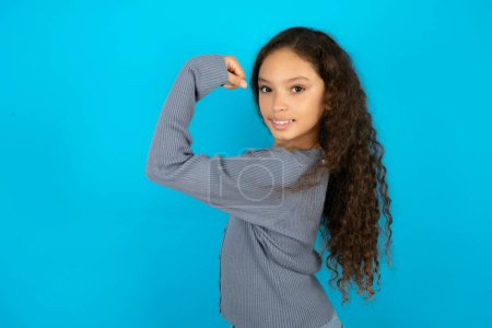 Photo for Portrait of powerful cheerful Beautiful kid girl wearing casual jacket over blue background showing muscles. - Royalty Free Image