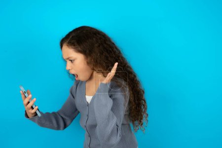 Foto de Angry Hermosa niña con chaqueta casual sobre fondo azul gritando por teléfono, teniendo una discusión con un empleado. Problemas en el trabajo. - Imagen libre de derechos