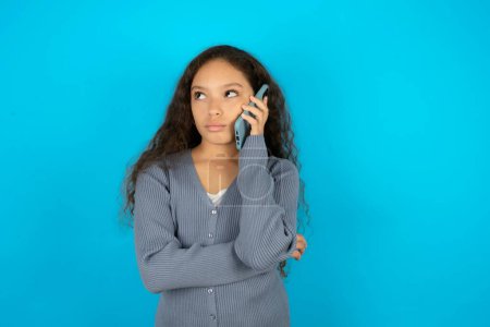 Photo for Sad Beautiful kid girl wearing casual jacket over blue background talking on smartphone. Communication concept. - Royalty Free Image