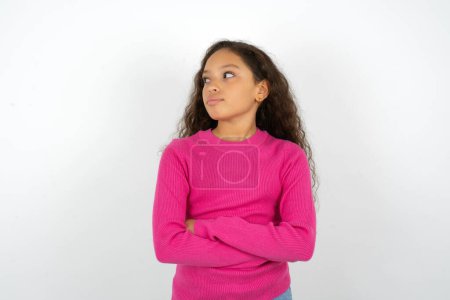 Photo for Charming thoughtful Beautiful kid girl wearing pink turtleneck over white background stands with arms folded concentrated somewhere with pensive expression thinks what to do - Royalty Free Image