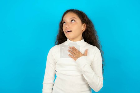 Photo for Joyful Beautiful kid girl wearing white turtleneck over blue background expresses positive emotions recalls something funny keeps hand on chest and giggles happily. - Royalty Free Image