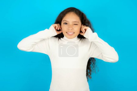 Photo for Happy Beautiful kid girl wearing white turtleneck over blue background ignores loud music and plugs ears with fingers asks to turn off sound - Royalty Free Image